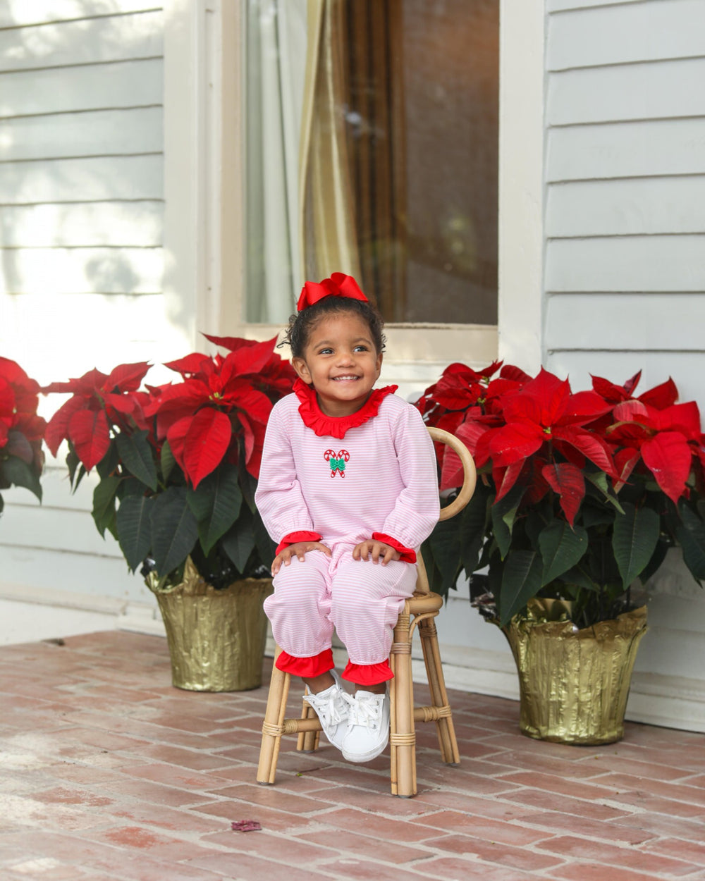 Child in pink Christmas-themed outfit sitting on a stool between potted poinsettias.