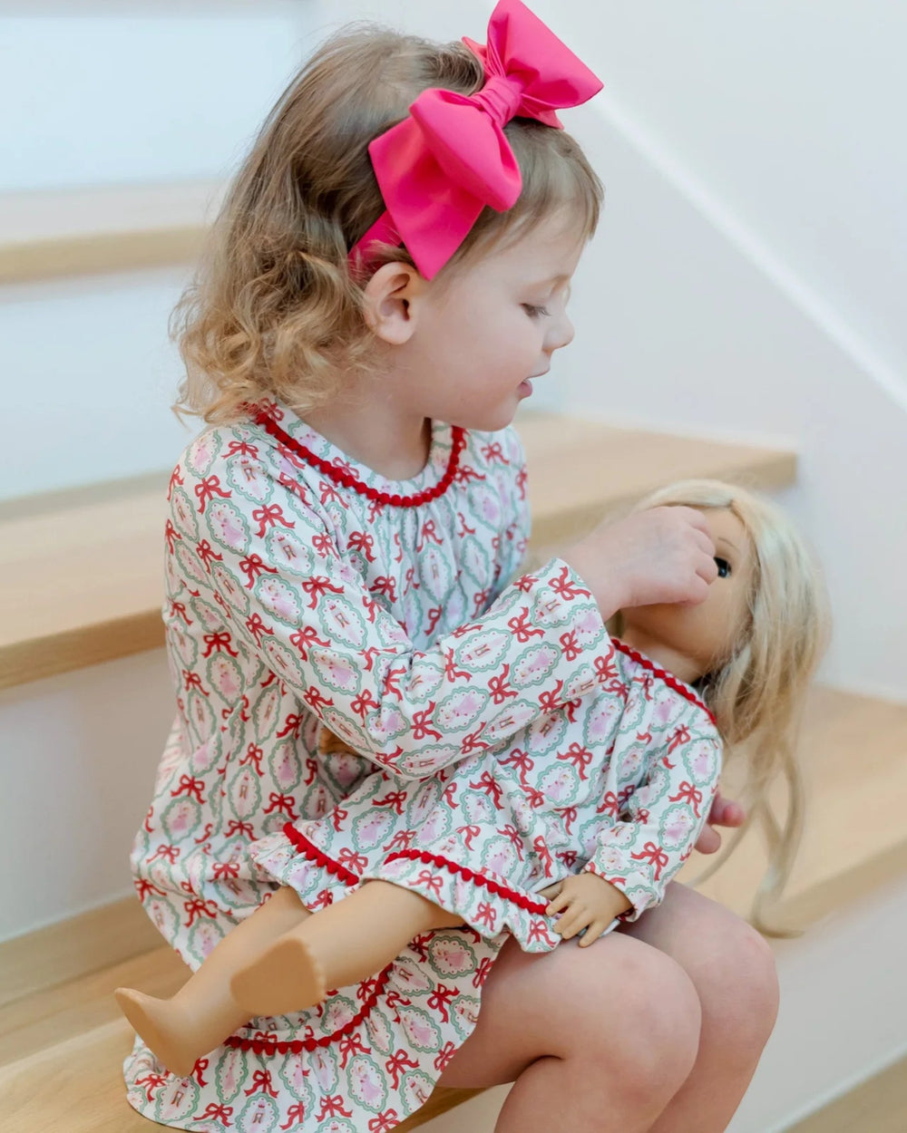 Child in a dress with red bows holding a doll on a wooden floor.