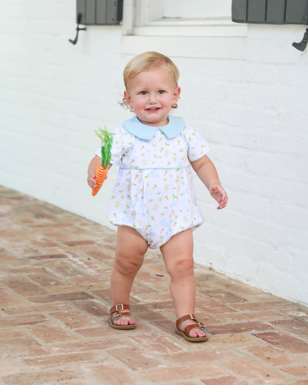 Child in a floral romper holding a toy carrot on a brick path.