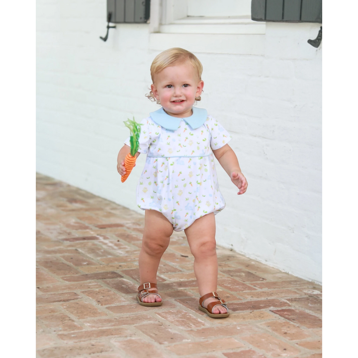 Child in a floral romper holding a toy carrot on a brick path.