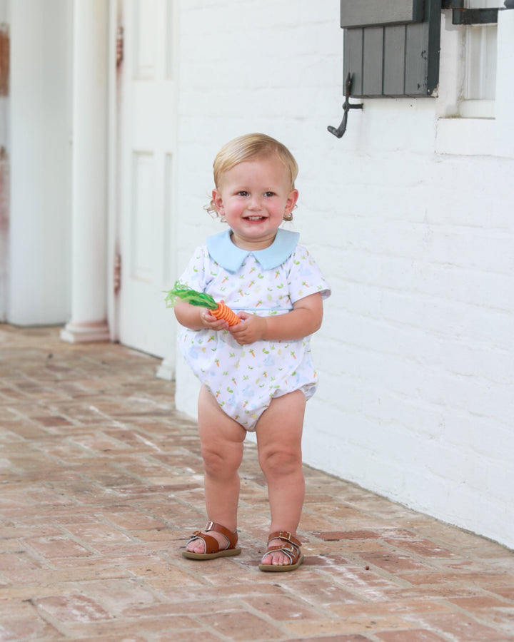 Child in a white onesie with blue collar holding carrots on a brick patio.