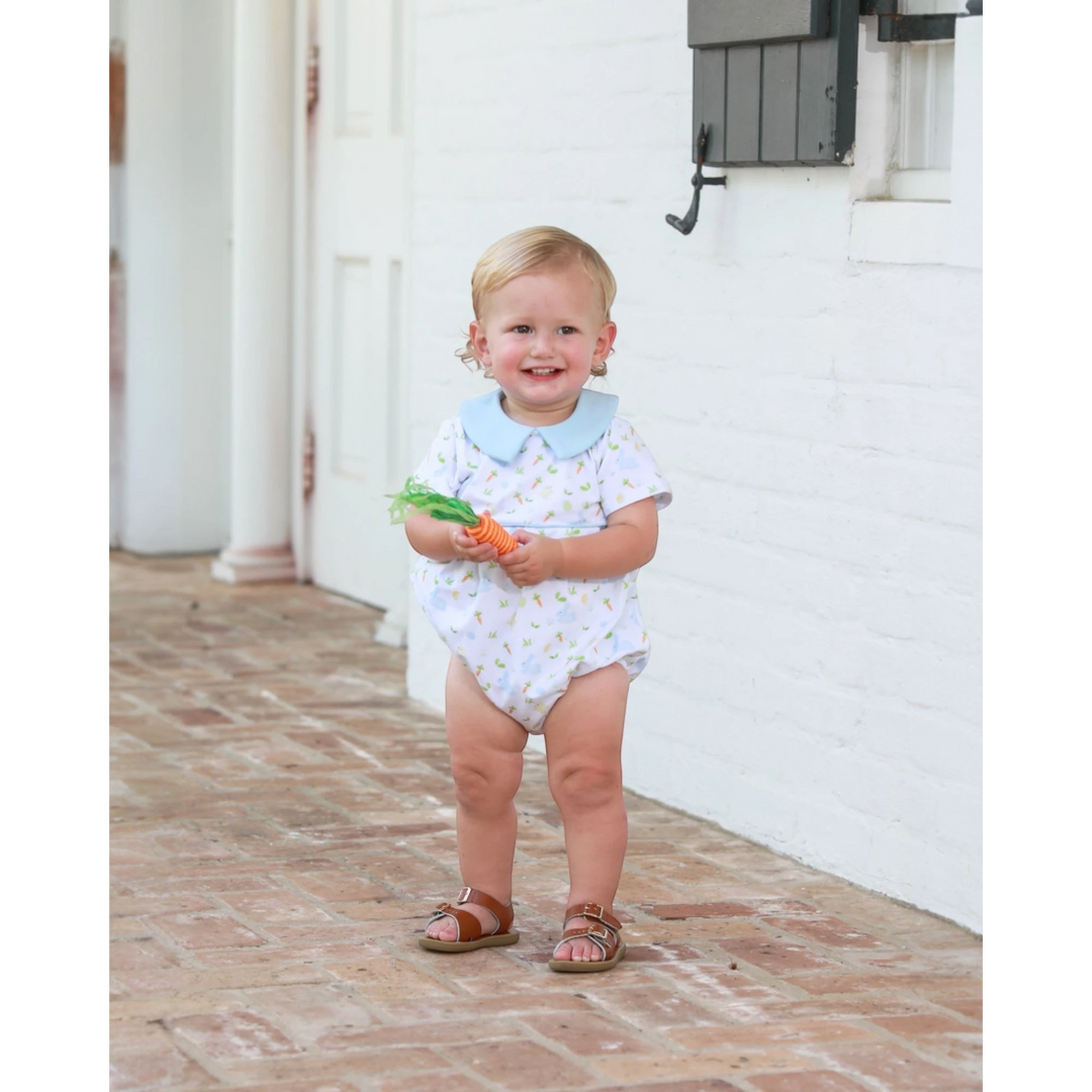 Child in a white onesie with blue collar holding carrots on a brick patio.