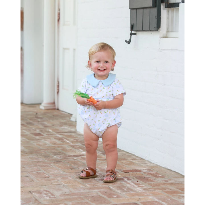 Child in a white onesie with blue collar holding carrots on a brick patio.