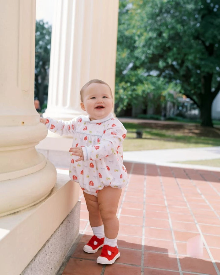 A baby standing outdoors next to a large cream-colored column, wearing a white long-sleeve romper with a red strawberry print, white socks, and red shoes, that store