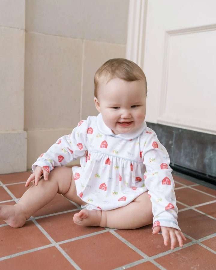 A baby sitting on a terracotta tiled floor wearing a white long-sleeve smocked bodysuit with a red and yellow house pattern, that store