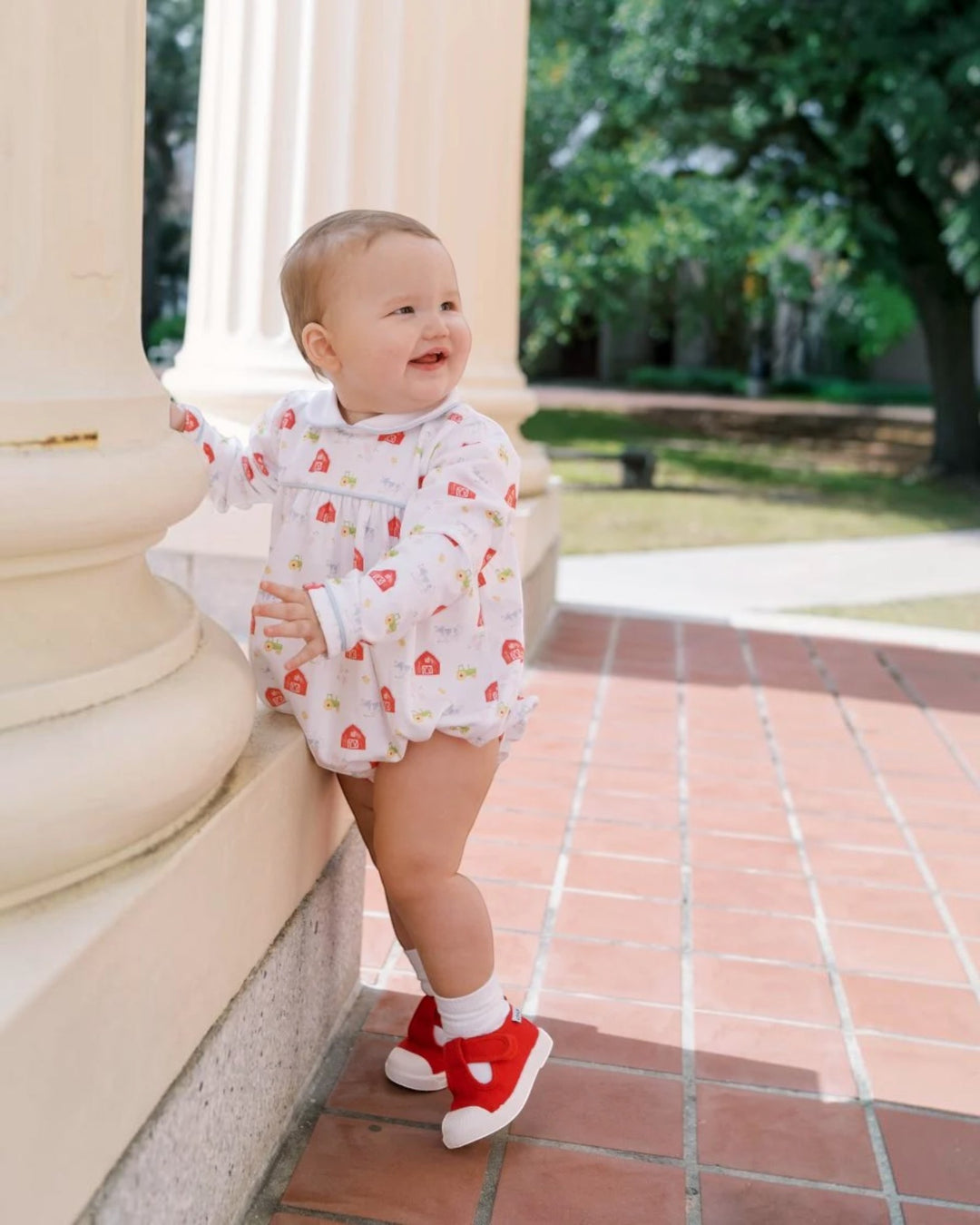 A baby standing outdoors next to a cream-colored stone column, wearing a white romper with a red strawberry print and red shoes, that store
