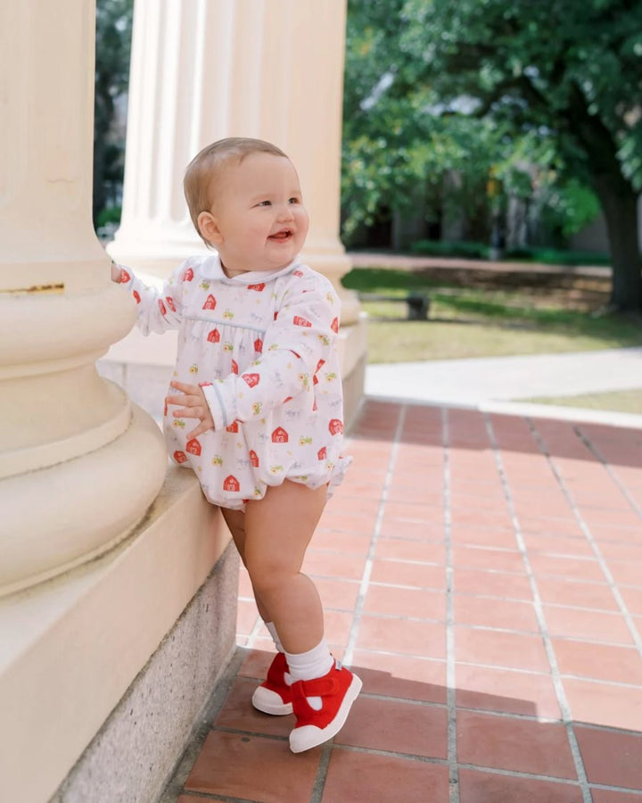 A baby standing outdoors next to a cream-colored stone column, wearing a white romper with a red strawberry print and red shoes, that store