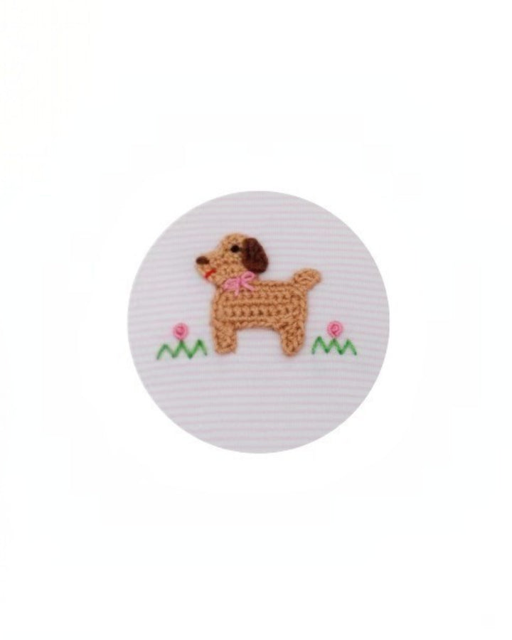 A circular white patch featuring a crocheted brown dog with a pink bow, green grass, and small pink flowers on a white striped fabric background, that store