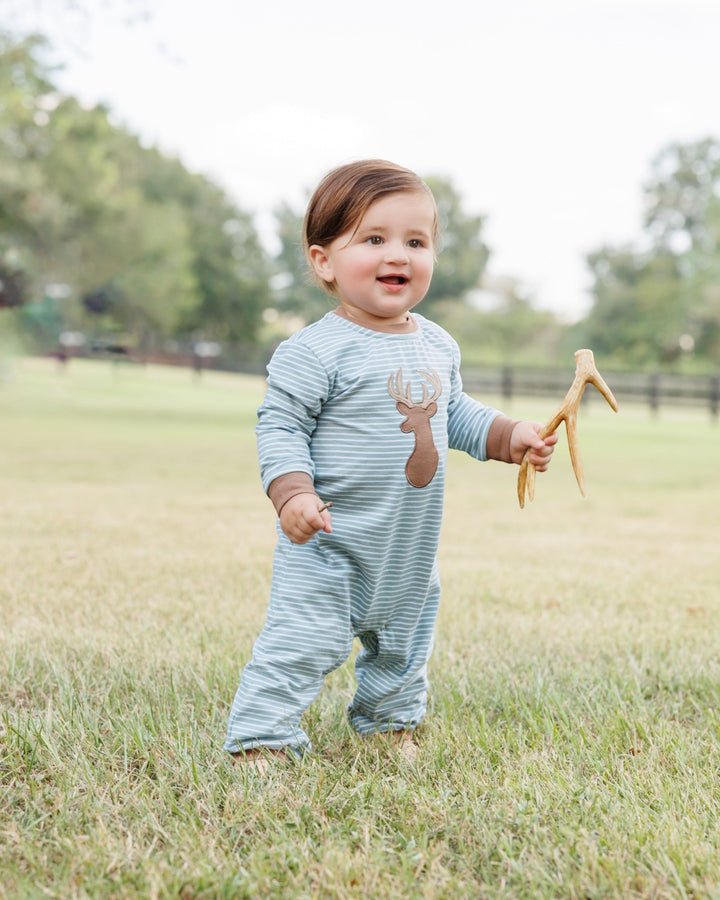 A baby wearing a light blue and white striped long-sleeve footless onesie with a brown deer graphic on the chest, standing in a grassy field and holding a small antler toy, that store