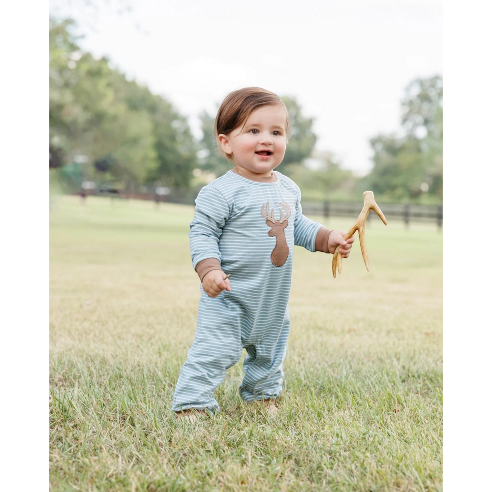 A baby wearing a light blue and white striped long-sleeve footless onesie with a brown deer graphic on the chest, standing in a grassy field and holding a small antler toy, that store
