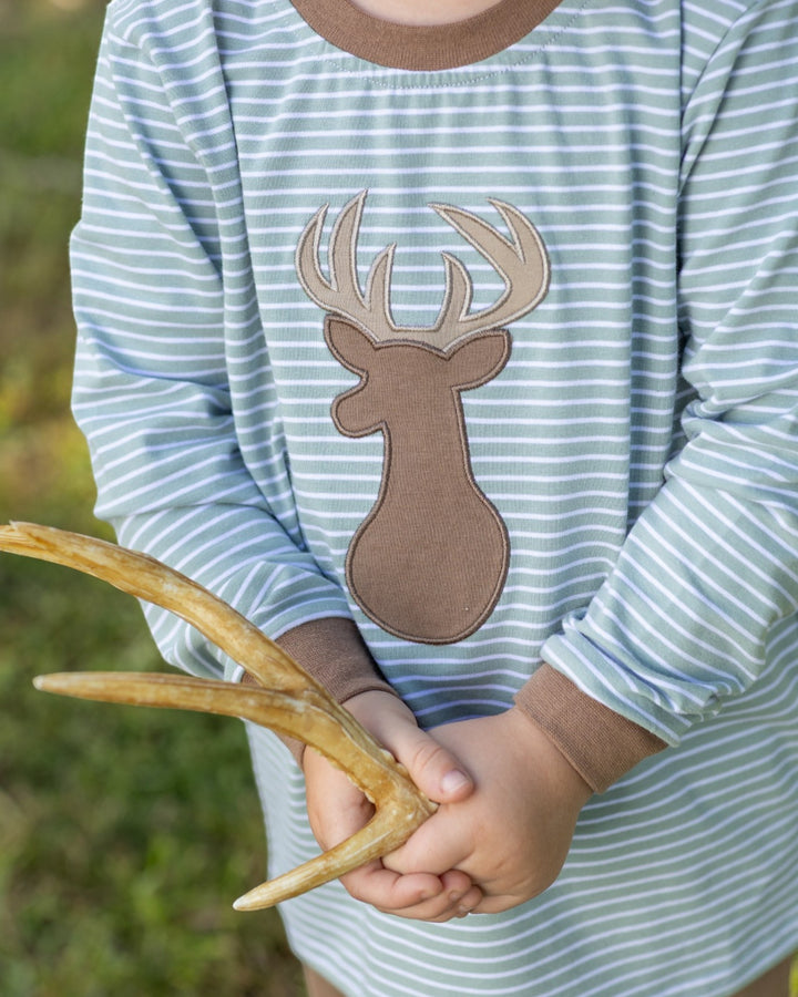 A close-up of a child wearing a light blue and white striped long-sleeve shirt with a brown deer applique on the chest, holding a small antler outdoors, that store