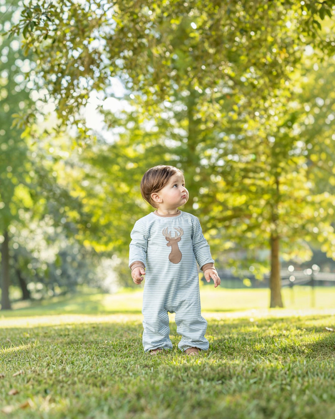 A baby standing on a grassy lawn under trees, wearing a light blue and white striped one-piece outfit with a brown reindeer graphic on the chest, that store