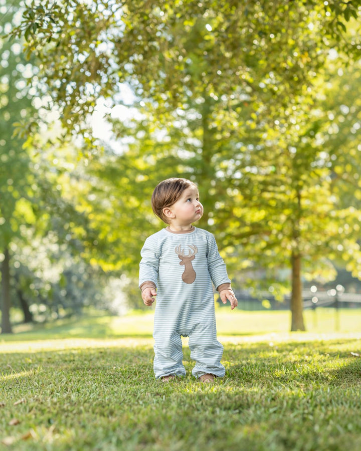 A baby standing on a grassy lawn under trees, wearing a light blue and white striped one-piece outfit with a brown reindeer graphic on the chest, that store