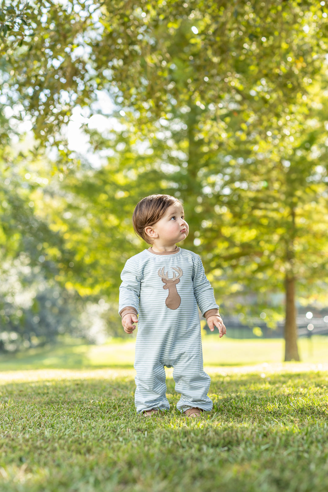 A toddler standing on grass in a park, wearing a light blue and white striped long-sleeve footless romper with a brown reindeer graphic on the chest, that store