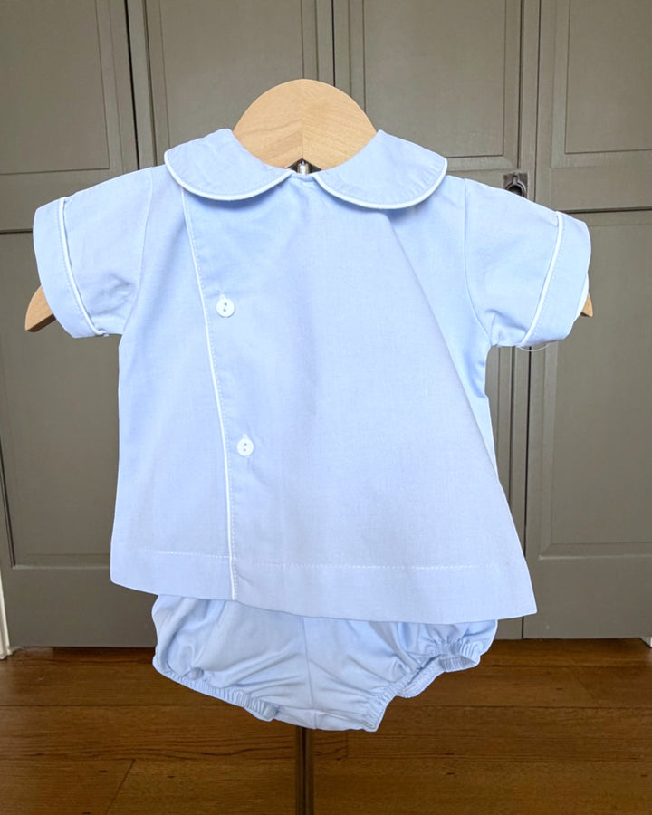 Light blue baby outfit with shirt and bloomers on a wooden hanger against a neutral background.