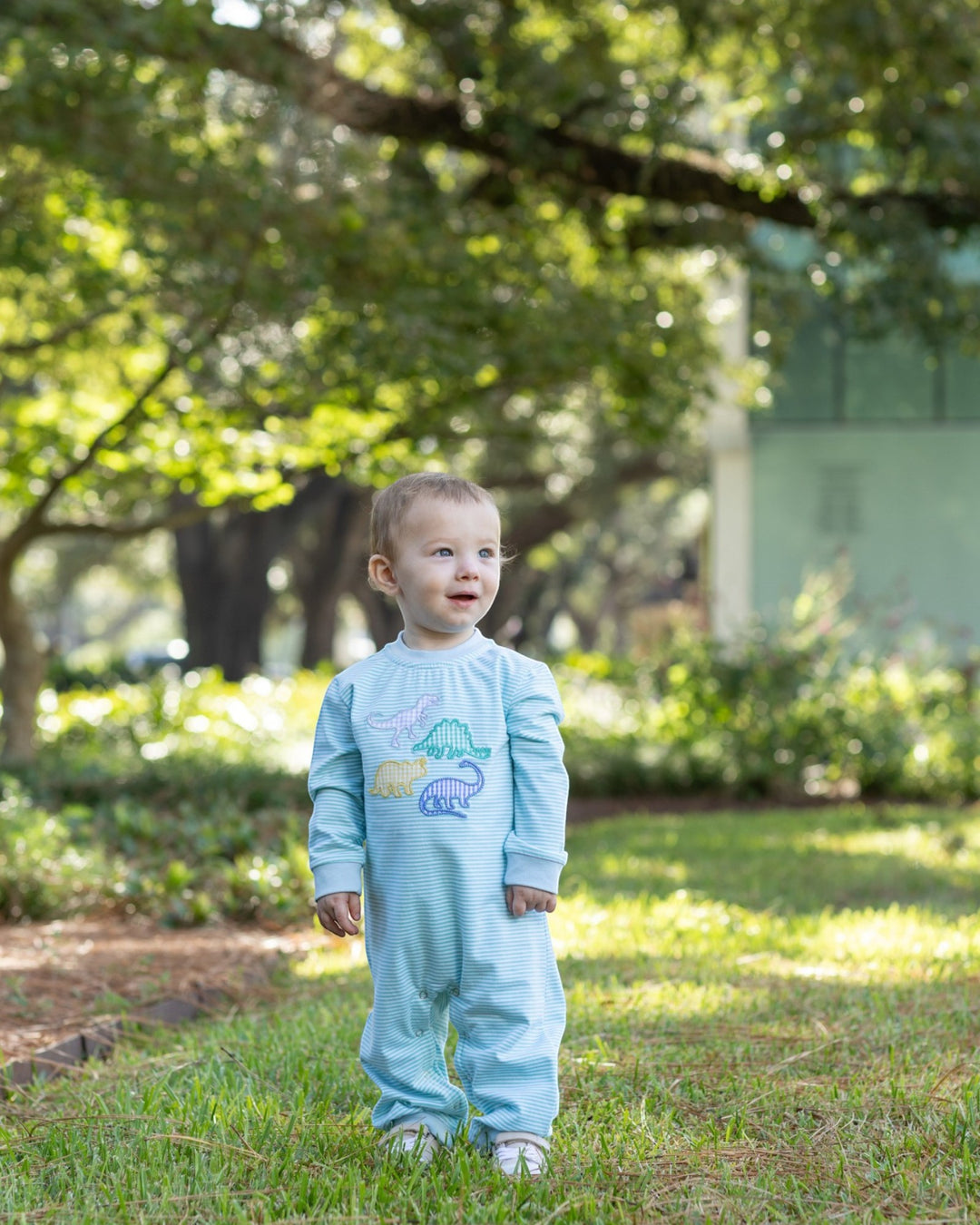A toddler standing outdoors on grass in front of trees and a building, wearing a light blue long-sleeve footed jumpsuit with a dinosaur graphic on the chest, that store