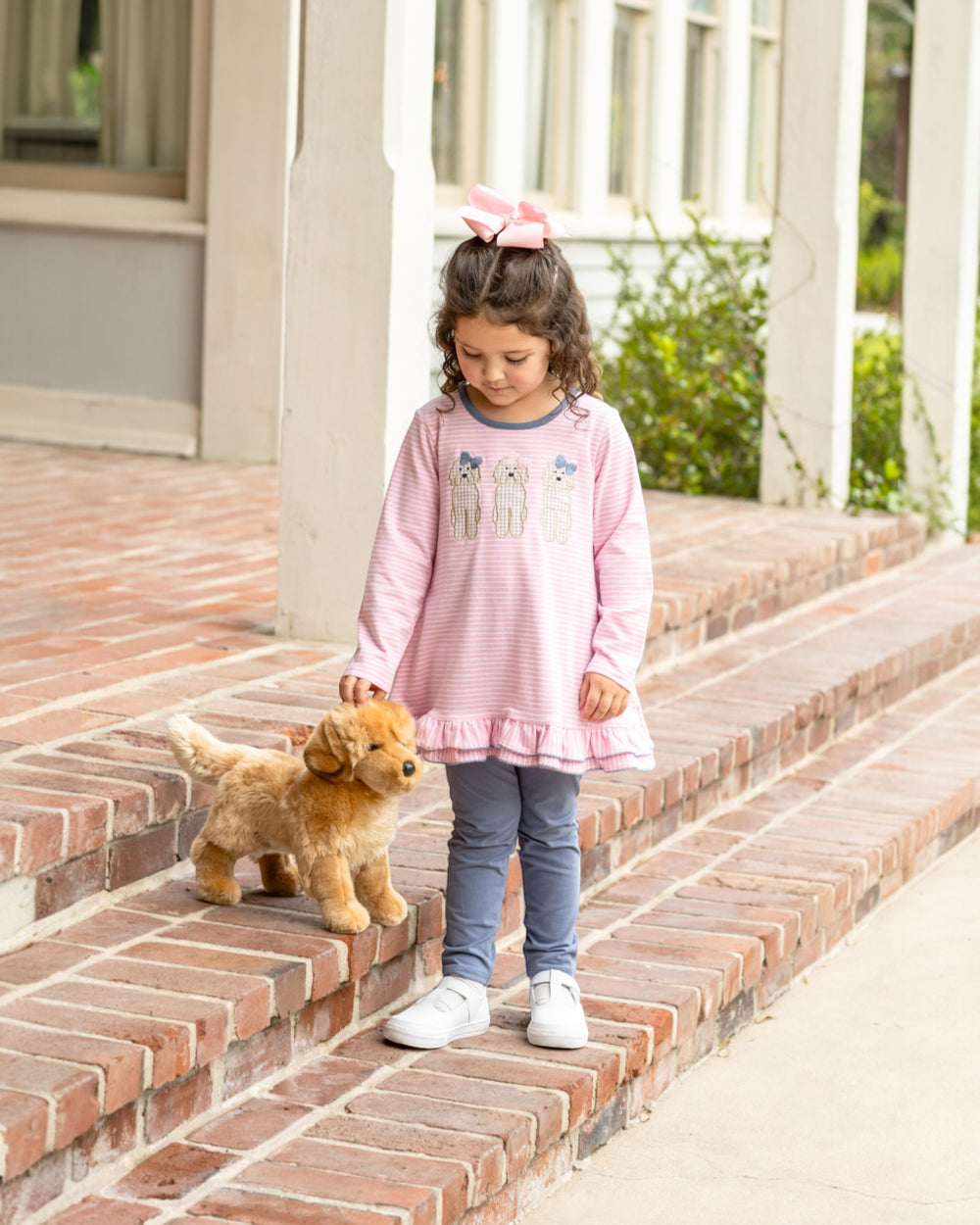 Young girl in a pink dress standing on brick steps holding a stuffed dog toy.