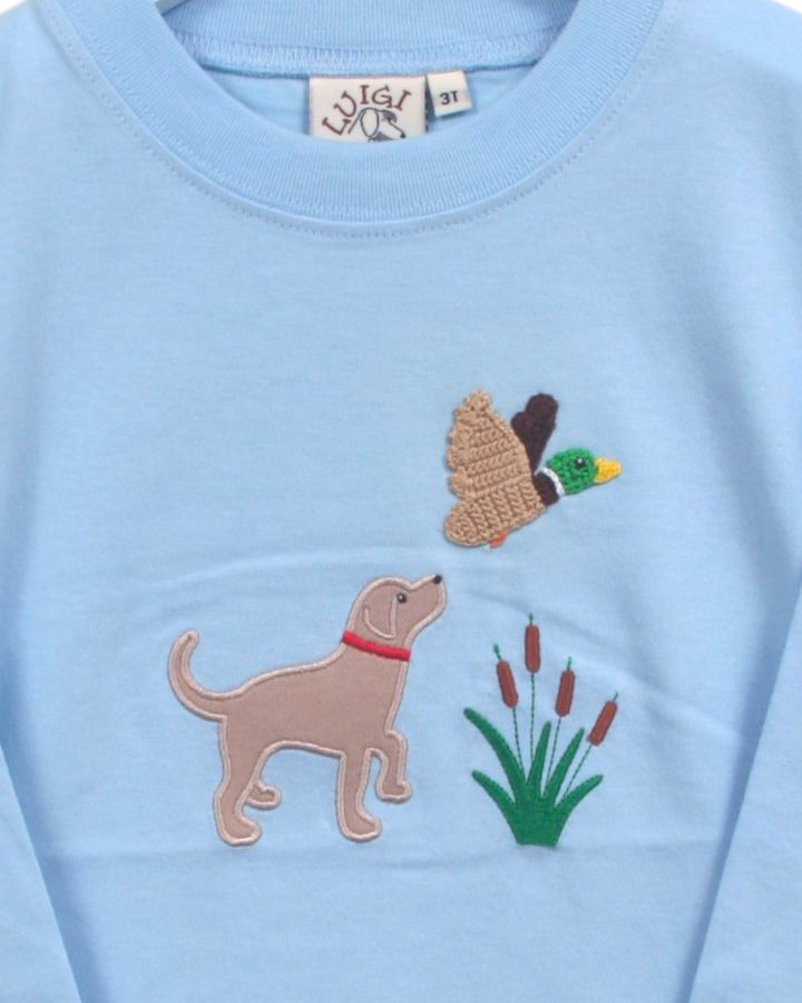 A close-up of a light blue long-sleeve shirt featuring embroidered dog and duck motifs with a small patch of cattails on the chest, that store