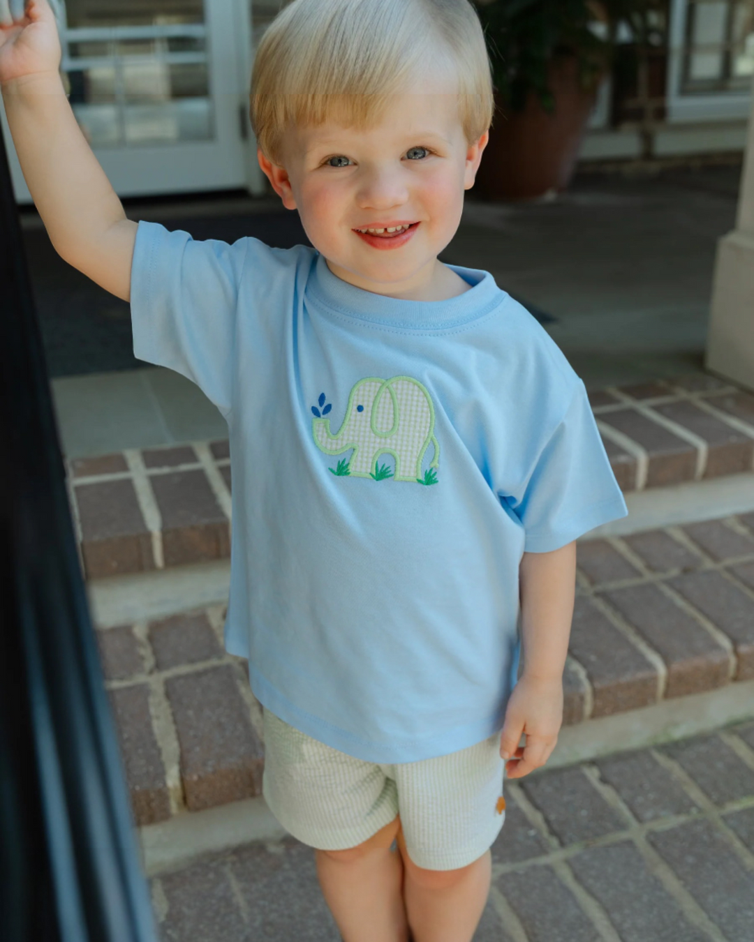 Child wearing a light blue shirt with an elephant design, standing on a porch.