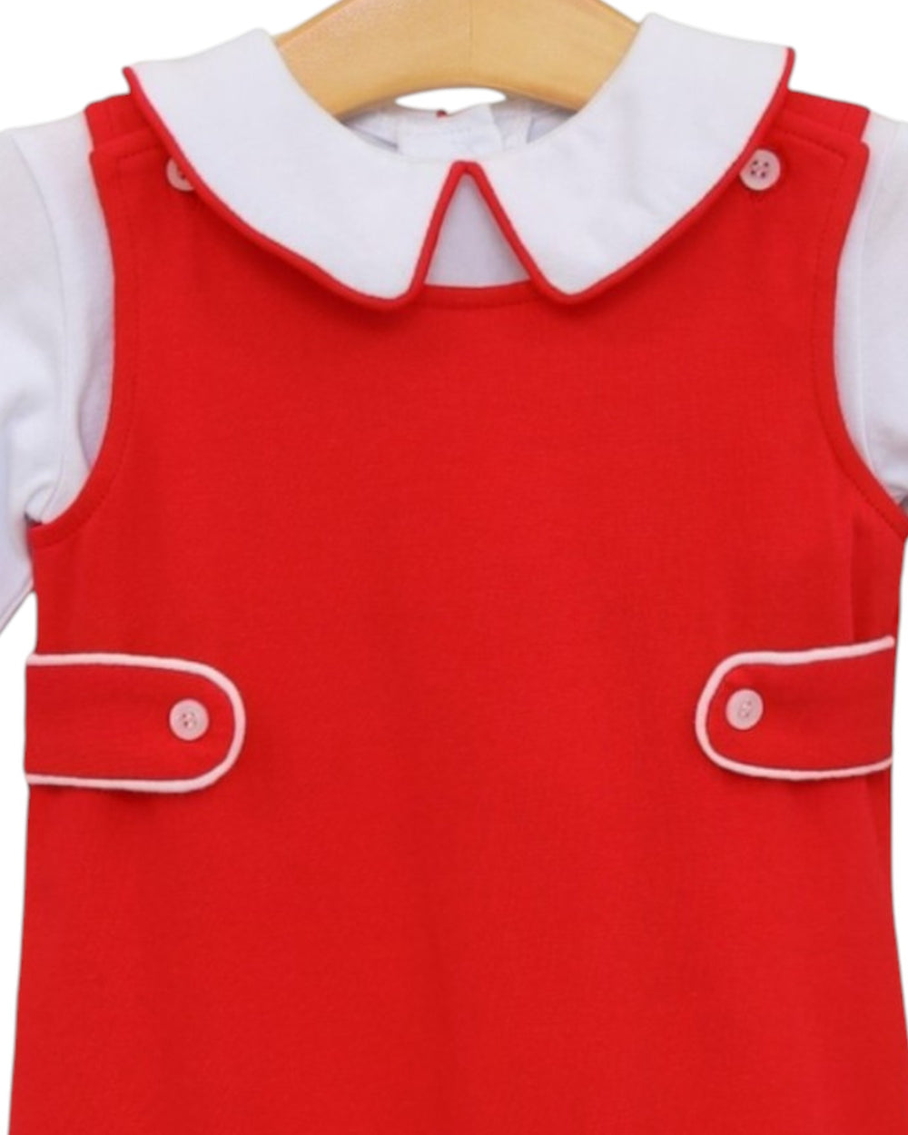 A red sleeveless dress with a white Peter Pan collar and white buttoned flap pockets, layered over a white short-sleeve top, shown on a wooden hanger against a white background, that store