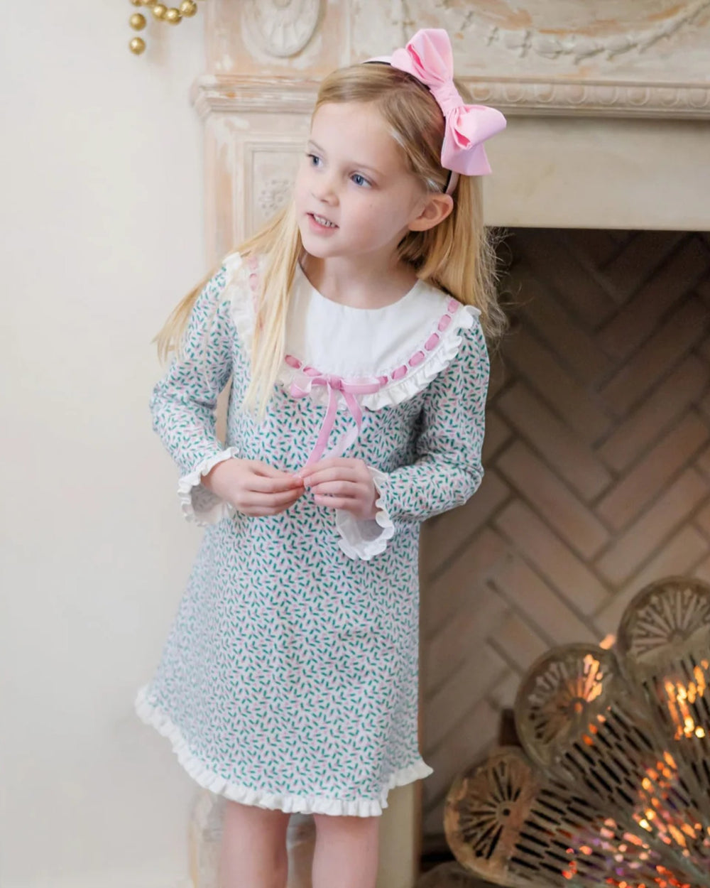 Young girl in a floral dress with a pink bow standing in front of a fireplace.