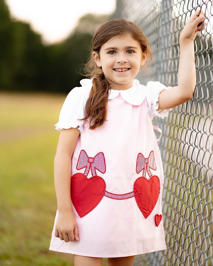 Young girl wearing a pink dress with red heart and bow patterns, standing next to a wire fence.