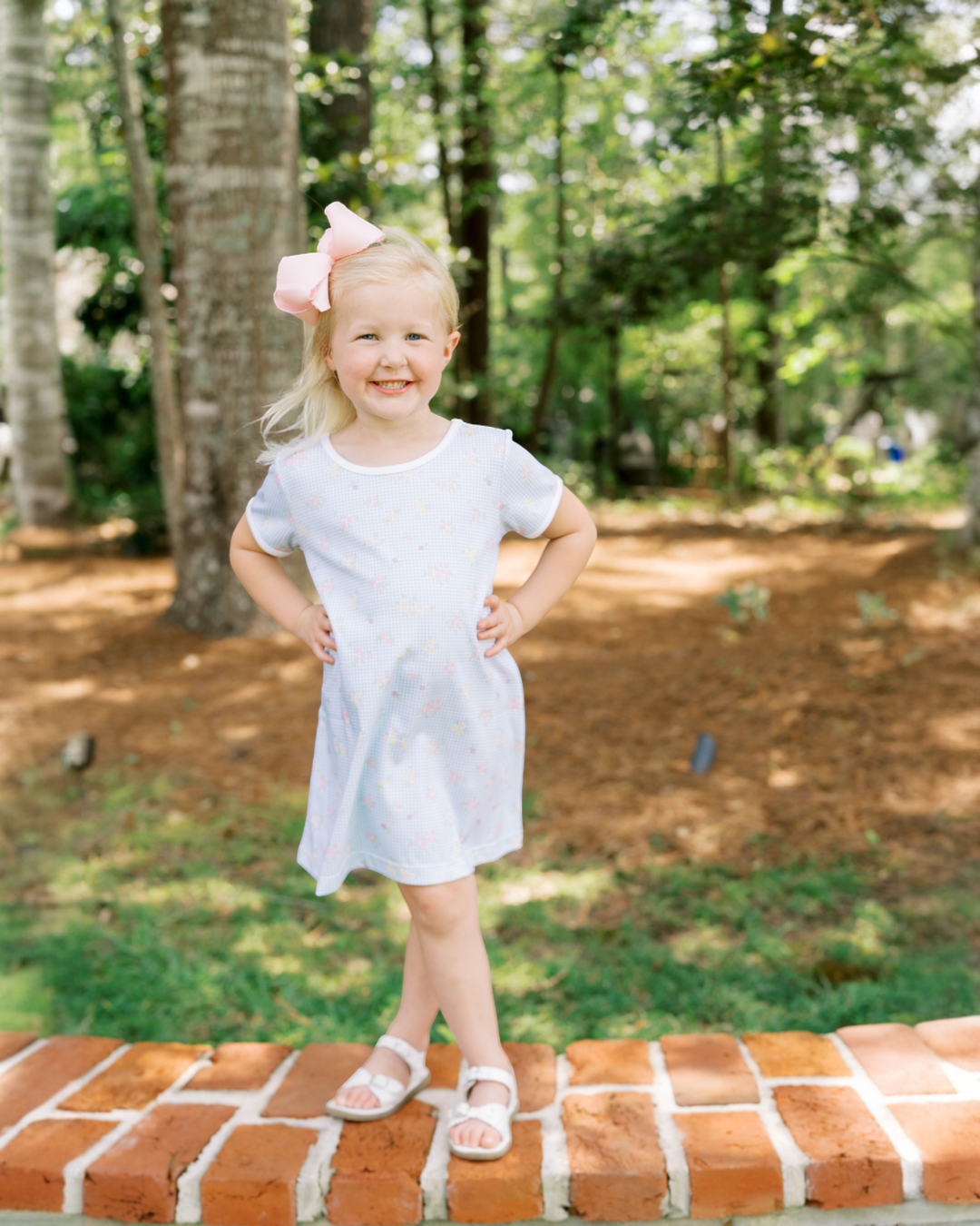 Young girl in a white dress with a pink bow standing on a brick wall outdoors.