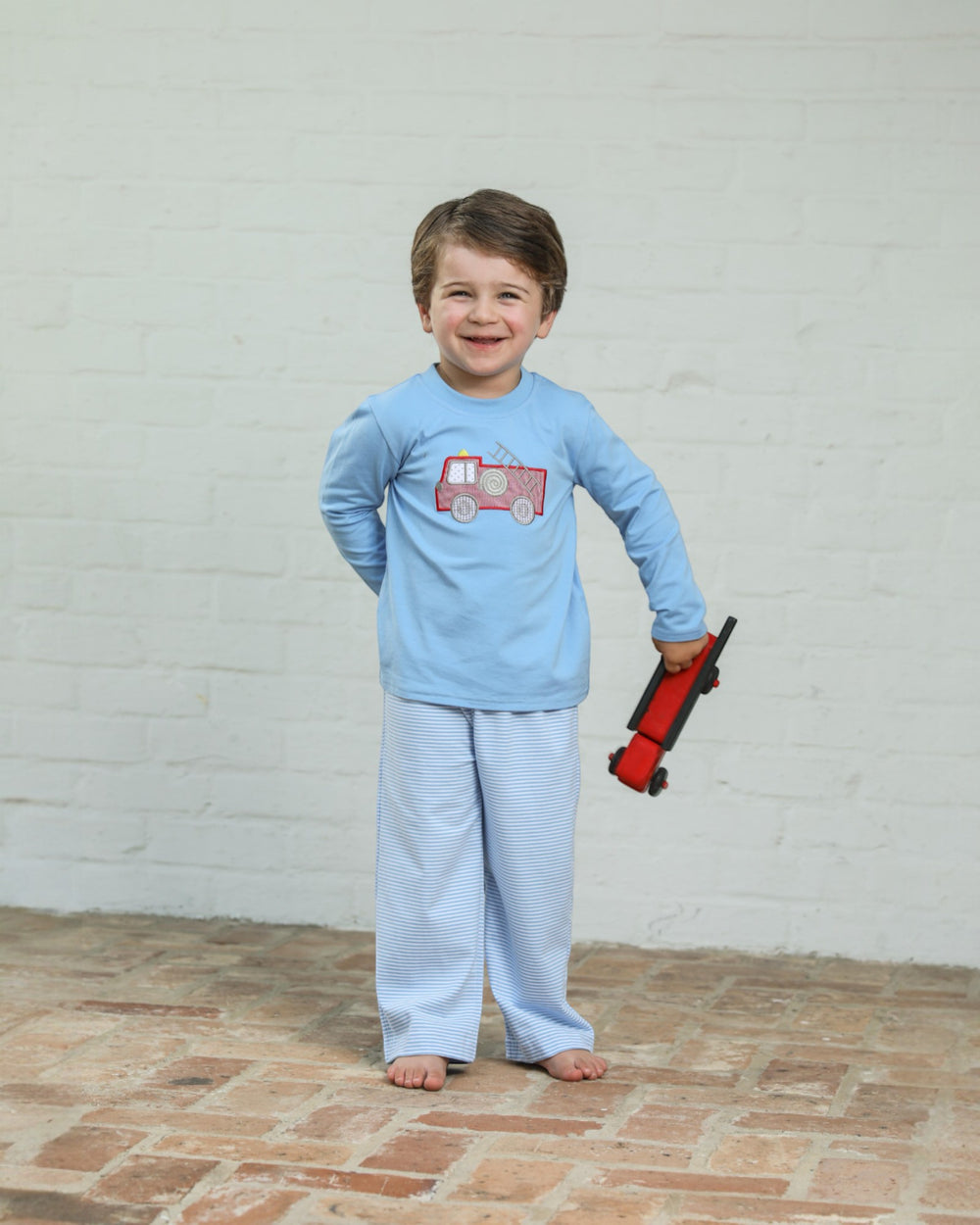 Child wearing a light blue pajama set with a red fire truck design, standing on a brick floor against a white brick wall.