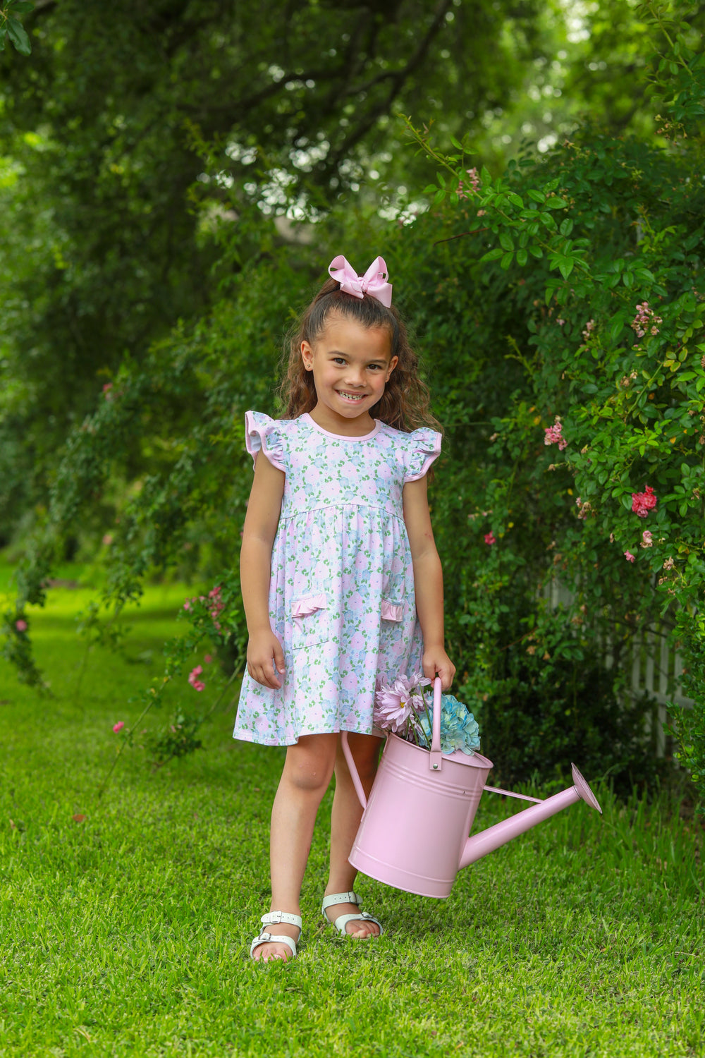 Young girl in a floral dress holding a pink watering can in a garden.