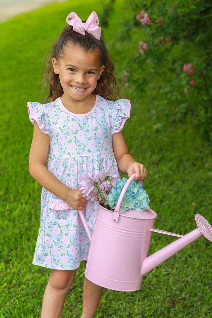 Young girl in a floral dress holding a pink watering can with flowers on a grassy background