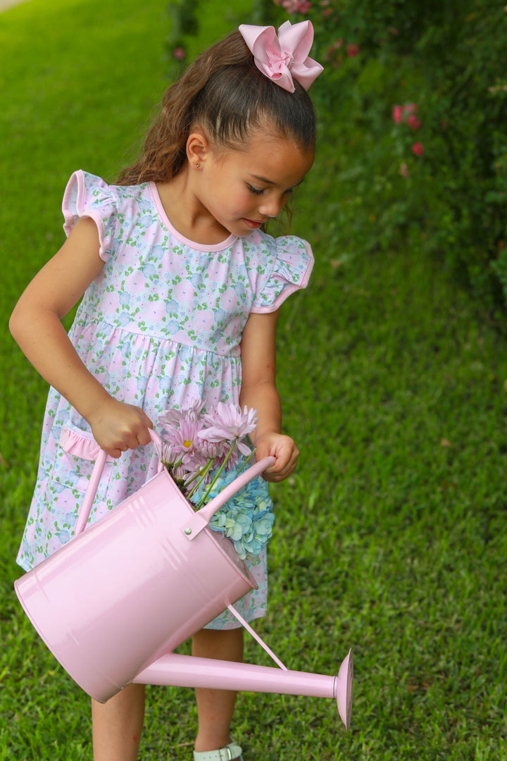 Young girl in a floral dress holding a pink watering can on grass