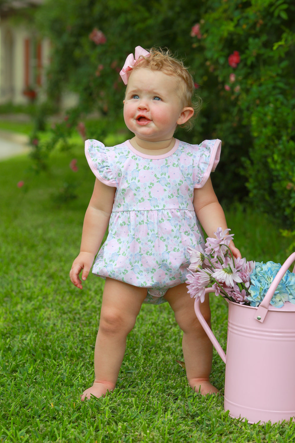 Child in a floral dress holding a pink watering can with flowers outdoors.