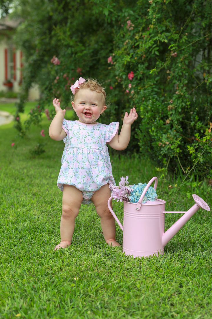 Child in a floral outfit standing next to a pink watering can with flowers in a garden setting