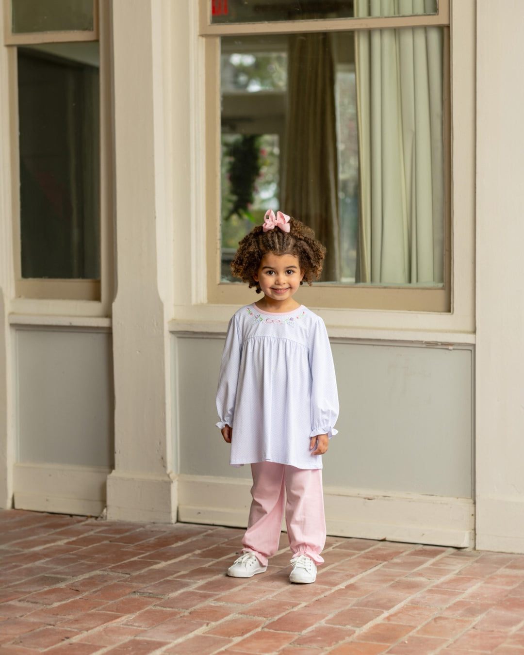 Young girl in a light purple dress and pink pants standing on a brick patio.
