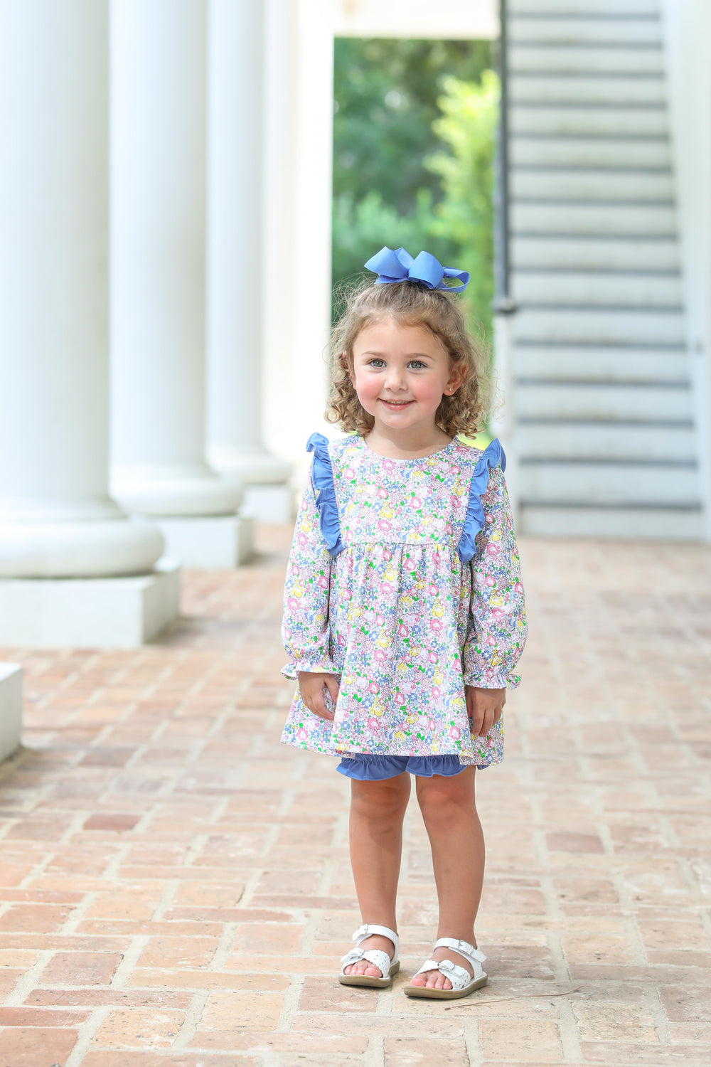 Young girl in a floral dress with blue ruffles standing on a brick path.