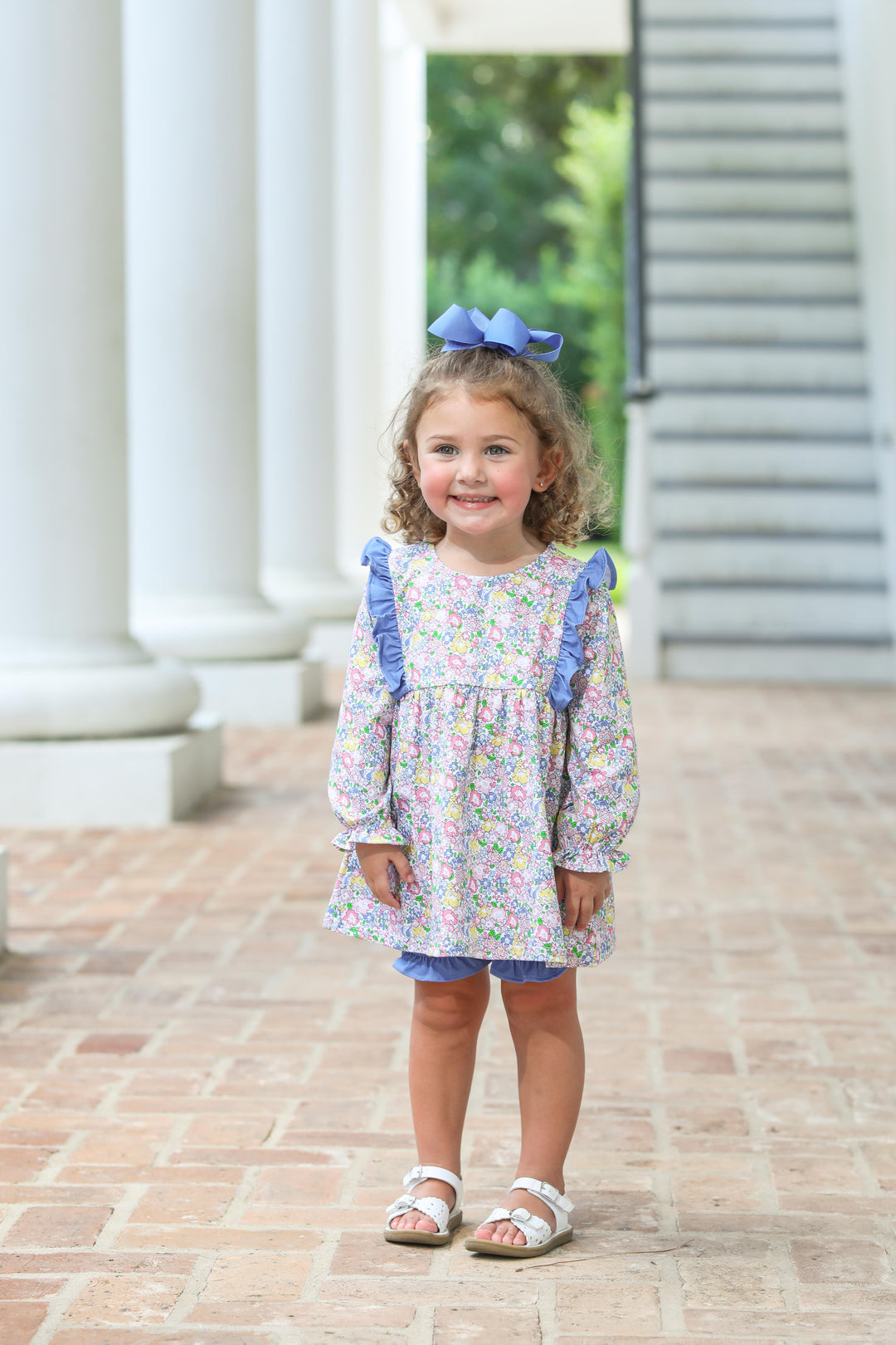 Young girl in a floral dress with blue ruffled sleeves standing on a brick path.