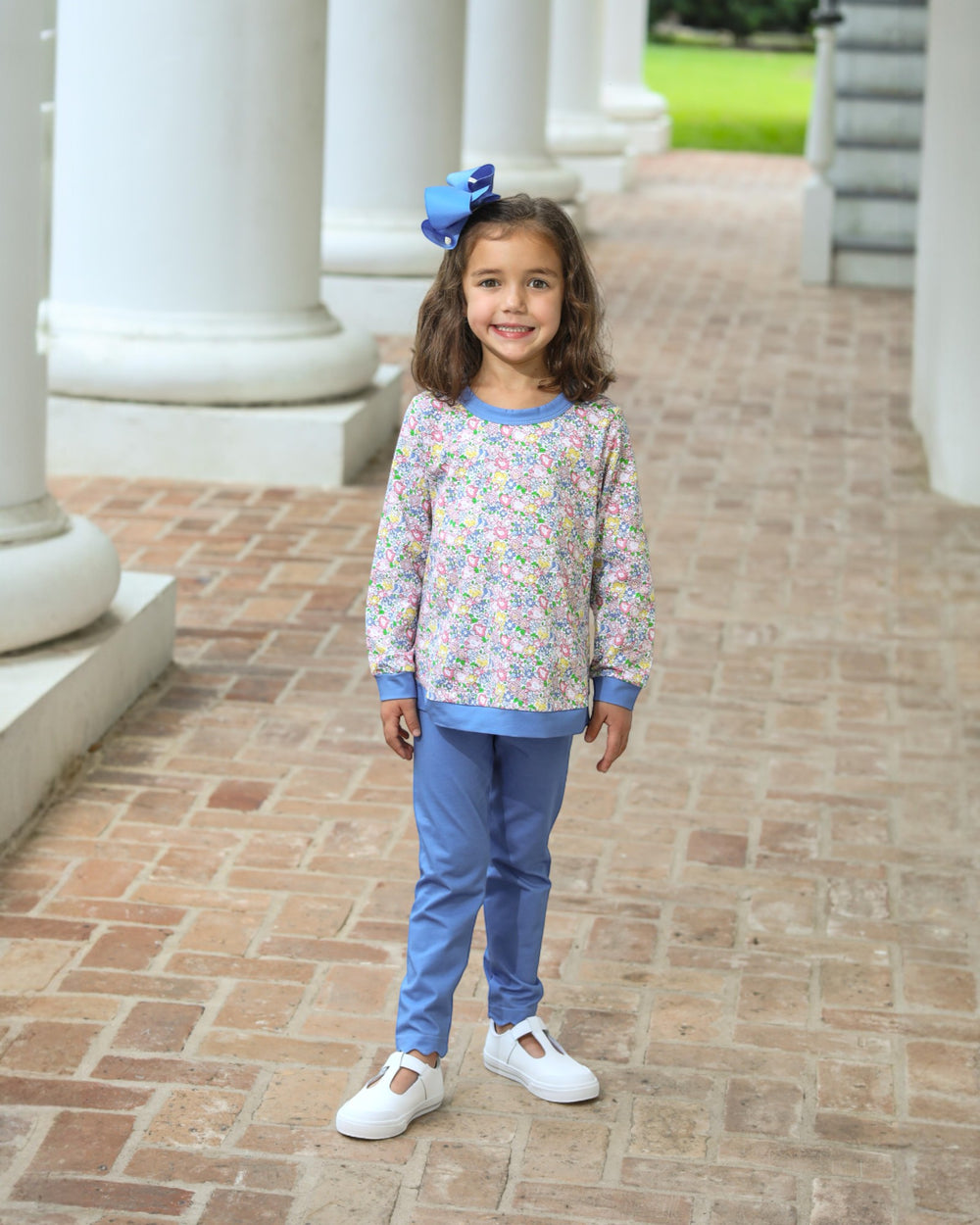 Young girl in a floral top and blue pants standing on a brick path with columns in the background