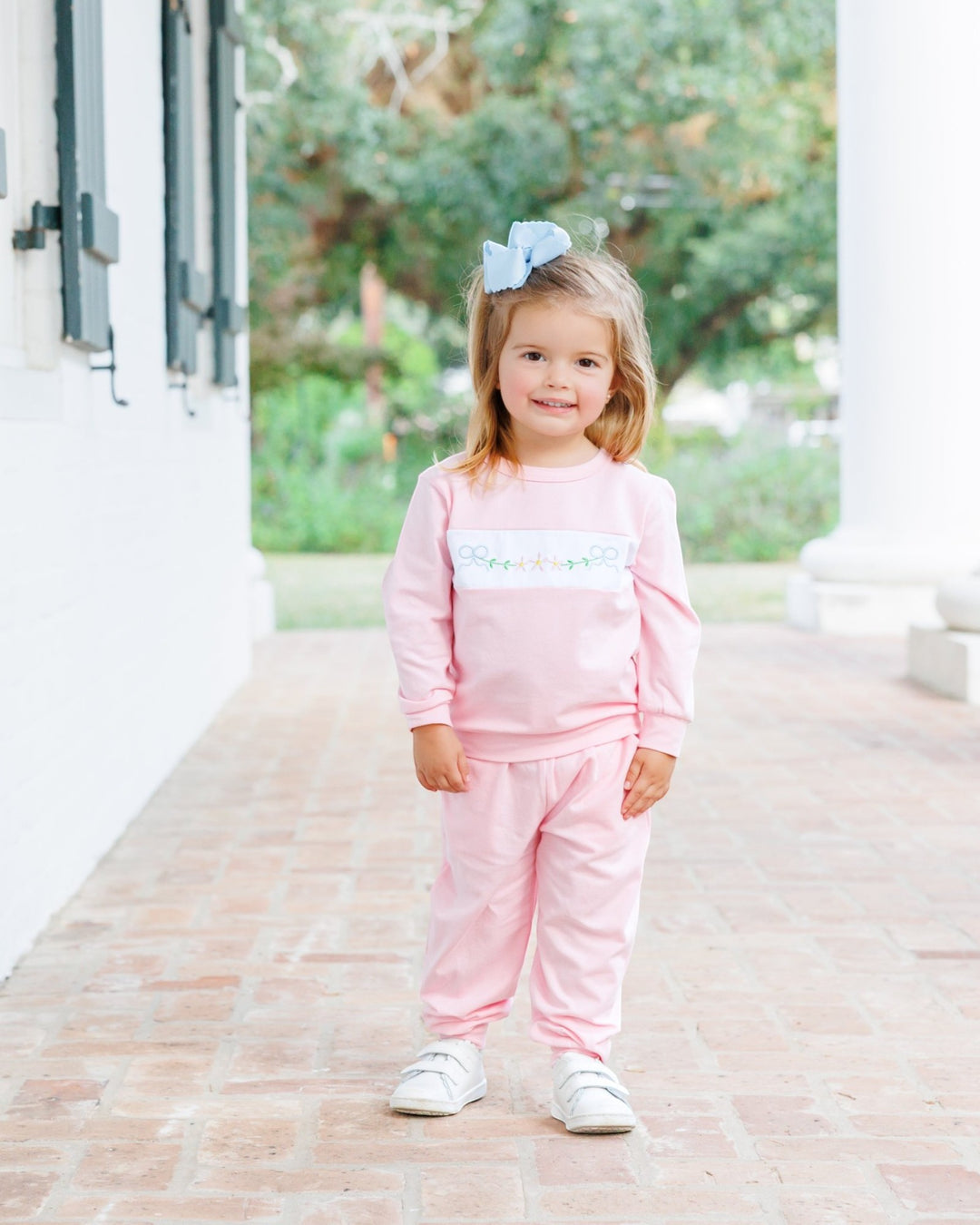 A young girl stands on a brick walkway wearing a light pink two-piece outfit with a white chest panel featuring small floral embroidery. She has a light blue bow in her hair and is wearing white shoes, that store