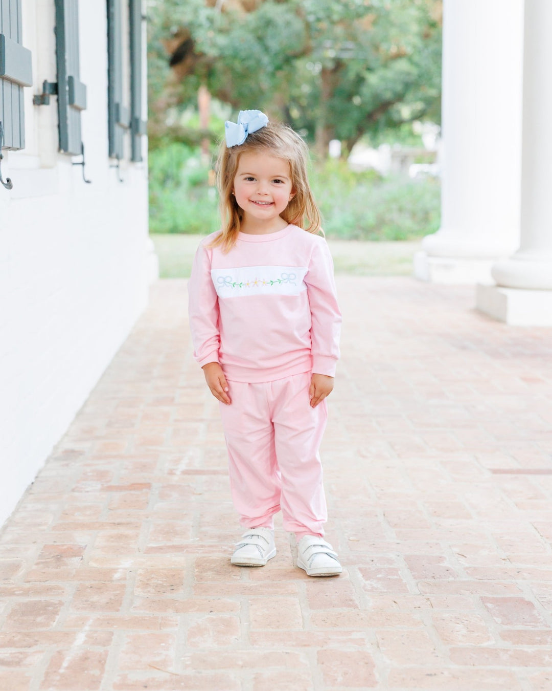 A young girl stands on a brick walkway wearing a light pink two-piece outfit with a white chest band featuring colorful script text, along with white sneakers and a light blue hair bow, that store