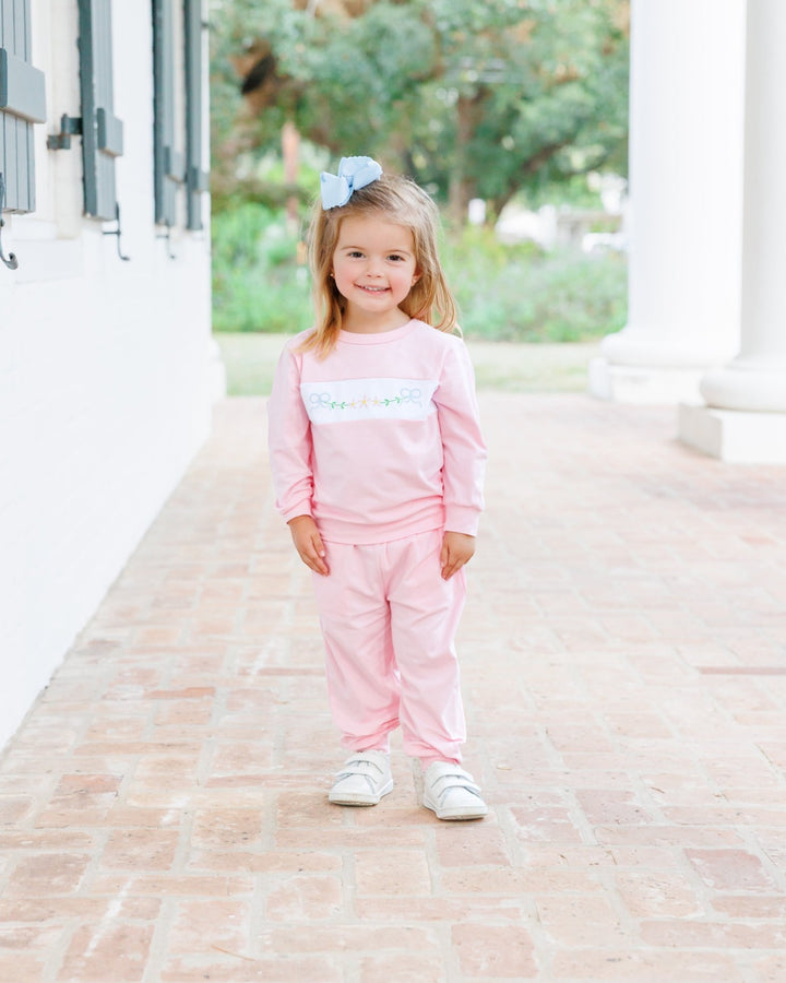 A young girl stands on a brick walkway wearing a light pink two-piece outfit with a white chest band featuring colorful script text, along with white sneakers and a light blue hair bow, that store