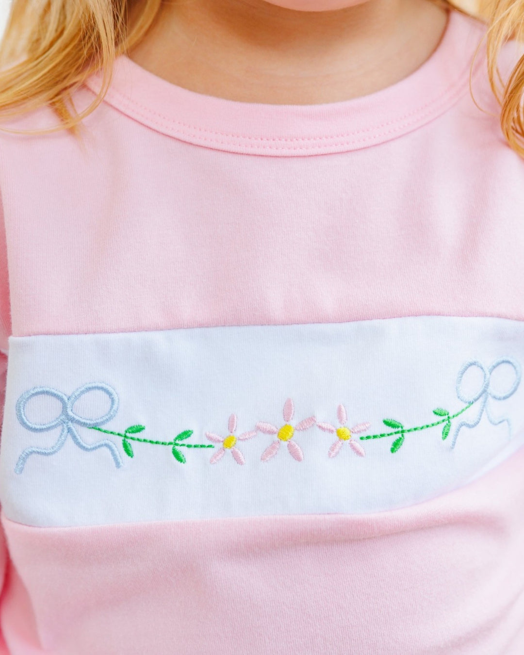 Close-up of a child wearing a pink long-sleeve top with a wide white horizontal band across the chest. The band features embroidered pink flowers with yellow centers, green stems and leaves, and blue bows on both sides, that store