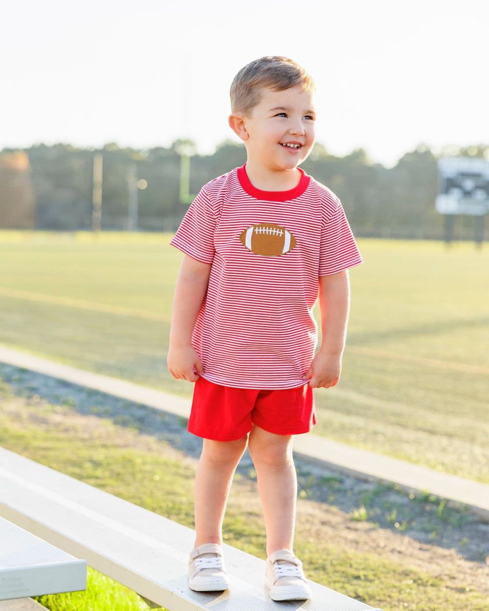 Child wearing a red and white striped shirt with a football design and red shorts, standing on a path outdoors.