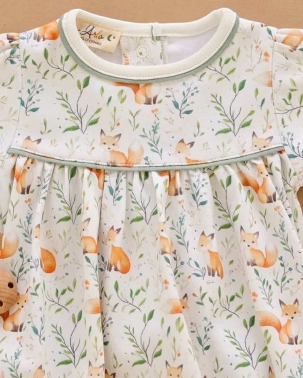 A close-up of a baby dress featuring an all-over print of orange foxes, green leaves, and delicate branches on a cream background, with a light sage green trim around the collar and waist, that store