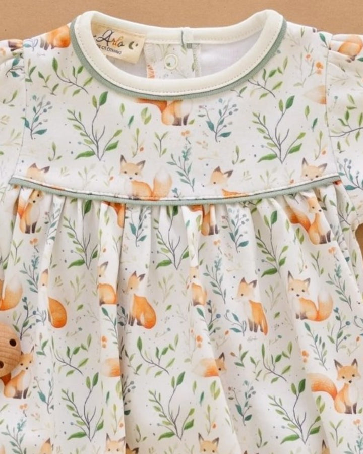 A close-up of a baby dress featuring an all-over print of orange foxes, green leaves, and delicate branches on a cream background, with a light sage green trim around the collar and waist, that store