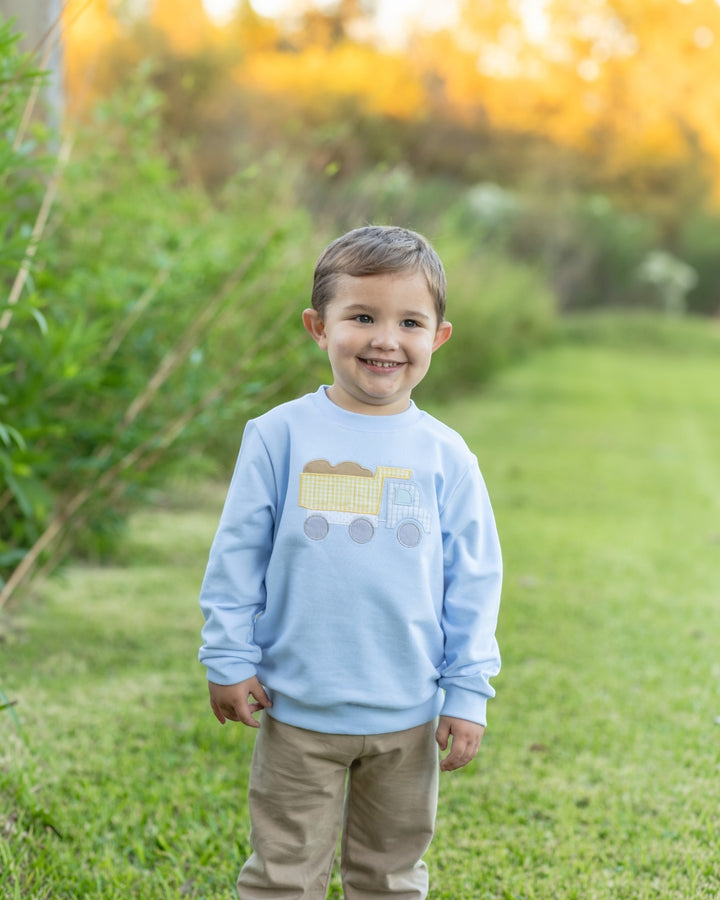 A young boy smiling outdoors on a grassy lawn at sunset, wearing a light blue crewneck sweatshirt with a yellow dump truck applique on the chest and tan pants, that store