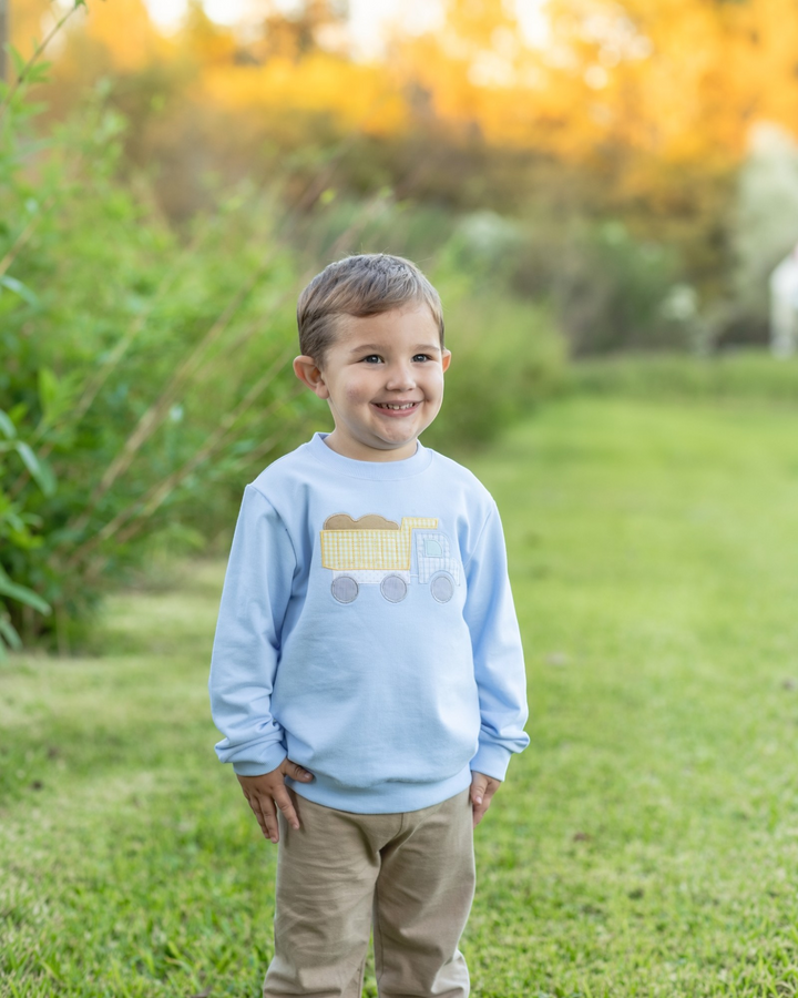 A young boy smiling outdoors on a grassy lawn, wearing a light blue crewneck sweatshirt with a yellow and gray train graphic on the front, paired with khaki pants, that store