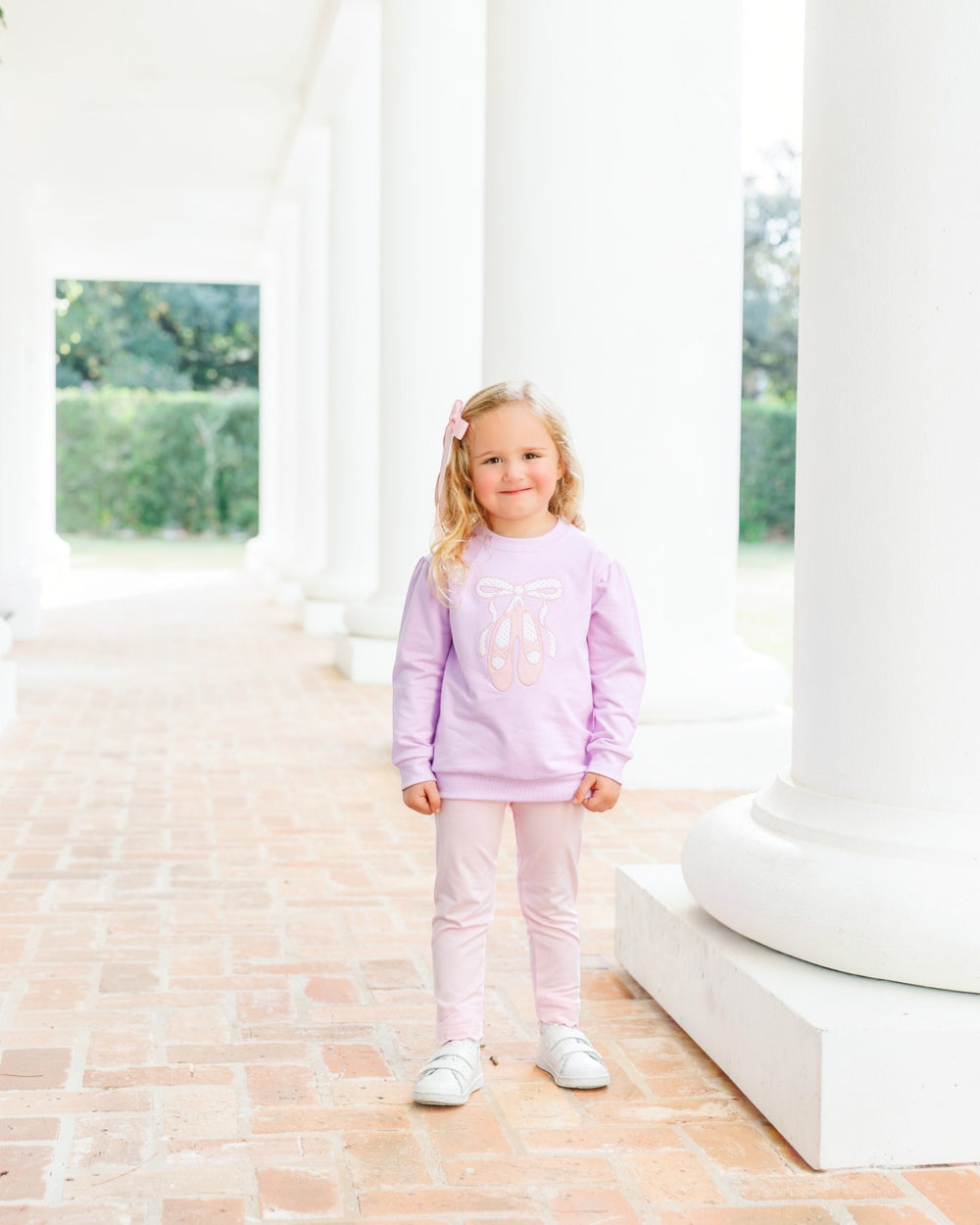 A young girl stands on a brick walkway between large white columns, wearing a light lavender long-sleeve sweatshirt with a bow graphic and light pink leggings, that store