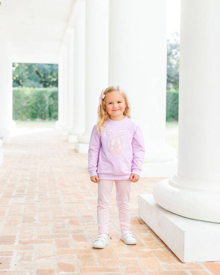 A young girl stands on a brick walkway between large white columns, wearing a light lavender long-sleeve sweatshirt with a bow graphic and light pink leggings, that store