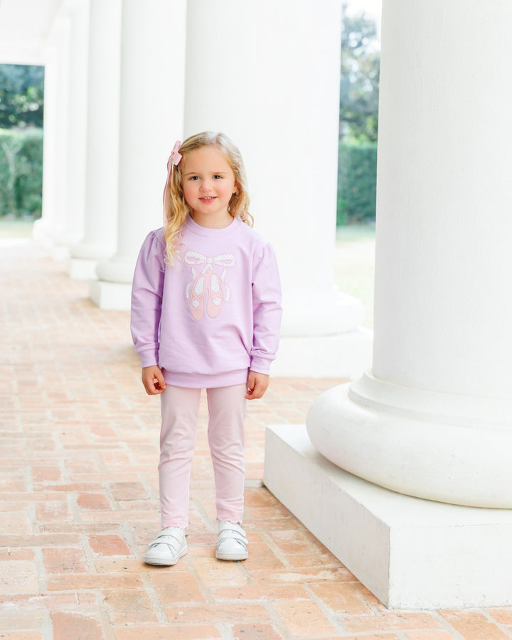 A young girl stands on a brick walkway between large white columns, wearing a light lavender sweatshirt with a pink bow graphic, light pink leggings, and white sneakers, that store