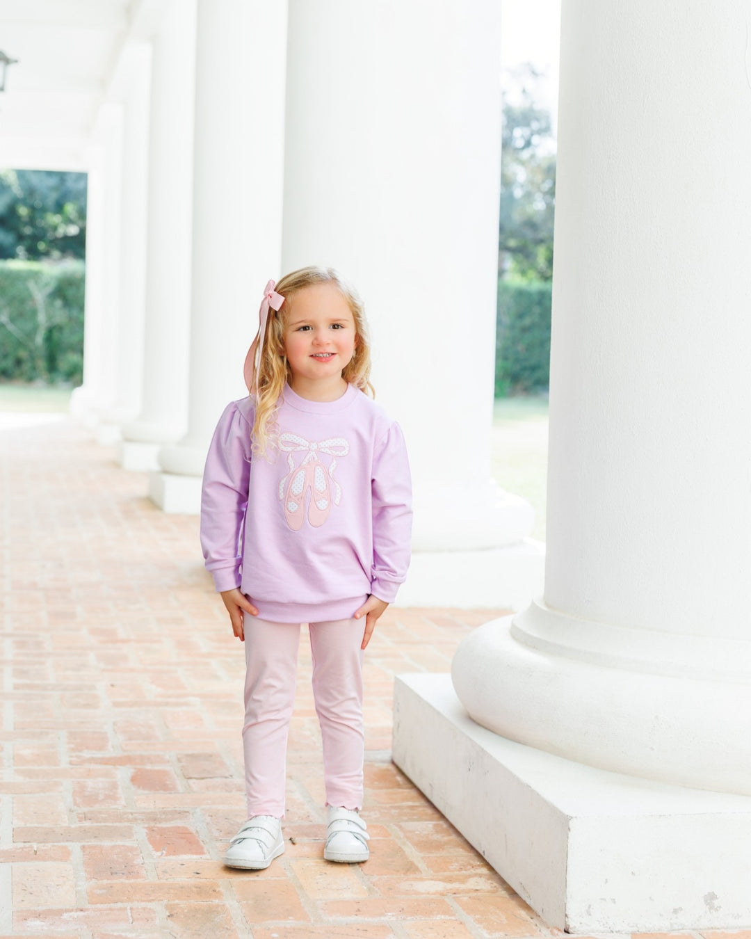 A young girl stands on a brick porch between large white columns, wearing a light lavender sweatshirt with a pink bow graphic and matching light pink leggings, that store